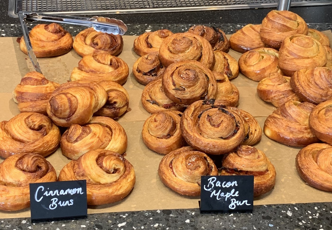 Bakery display of glazed pastries with chalkboard labels reading 'Cinnamon Buns' and 'Bacon Maple Bun'